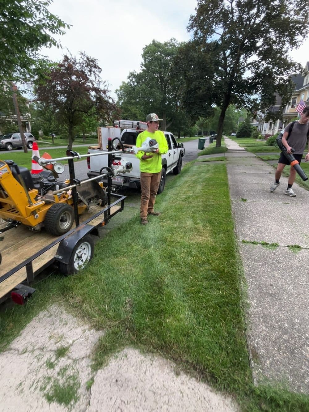 Lawn care professional adjusting equipment near a trailer on a residential street.
