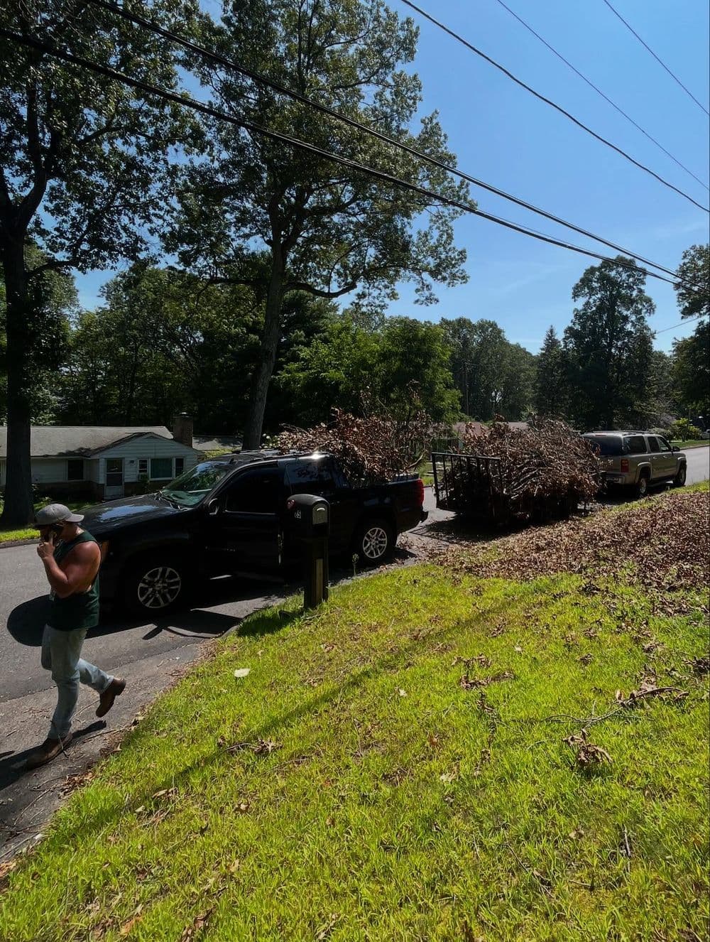 Man walking past parked SUVs with a trailer loaded with branches on a sunny day.
