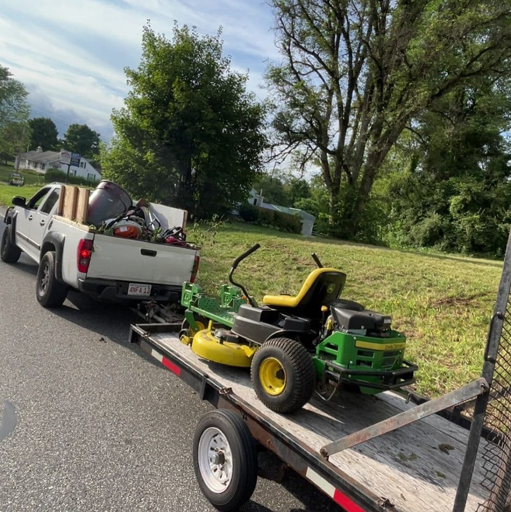 Pickup truck towing a trailer with a lawn mower and gardening tools on a grassy road.