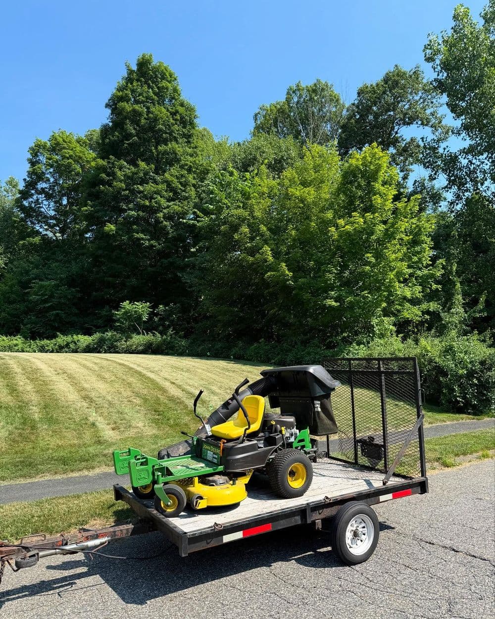 Zero-turn mower on trailer parked beside freshly mowed lawn and lush green trees.